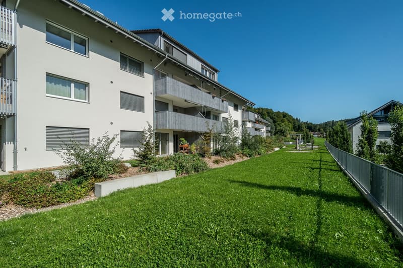Modern multi-story residential building with white exterior, multiple balconies, and surrounding green lawn.