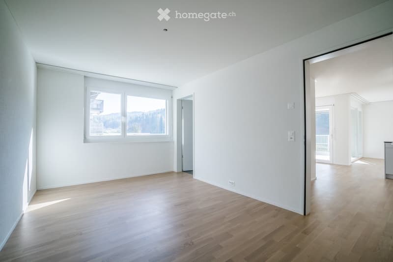 Empty room with light wooden flooring and white walls, featuring large windows and an open doorway leading to another space.