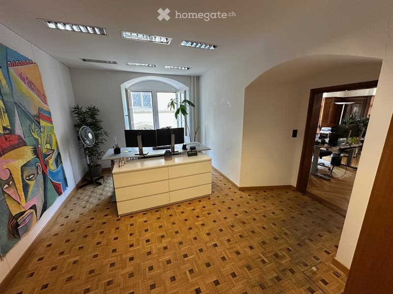 Office space with a reception desk, two computer screens, a fan, a potted plant, and a colorful mural on the wall. The floor is made of herringbone-patterned wood.