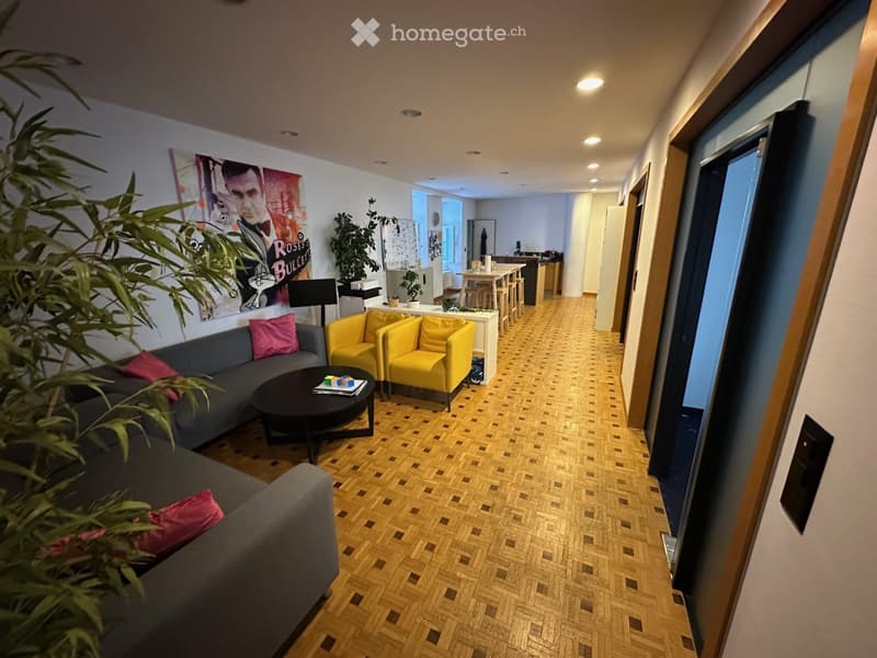 Living room with grey sofa, pink cushions, yellow chairs, coffee table, and a poster on the wall. The room has wooden flooring and leads to a kitchen area with a dining table and chairs.