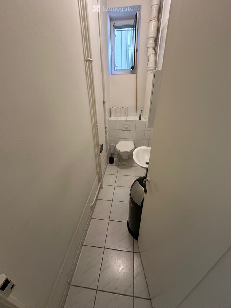 Small bathroom with white tiled floor and walls, toilet, sink, toilet paper, and a trash bin.