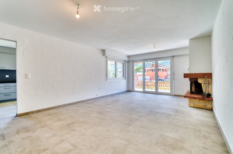 Empty living room with tiled floor, white textured walls, large sliding glass doors leading to outdoor area, fireplace with wooden mantel, and visible kitchen cabinets in the background.