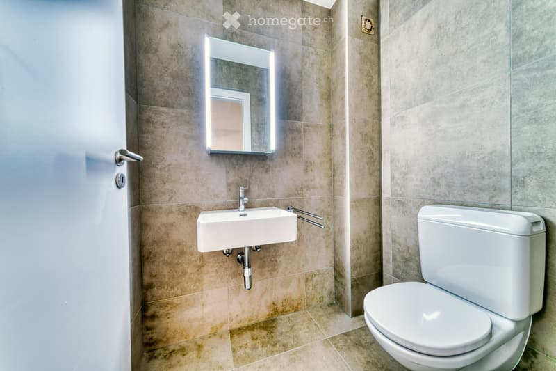 Modern bathroom with beige tiled walls and floor, white sink, toilet, and illuminated mirror above the sink.