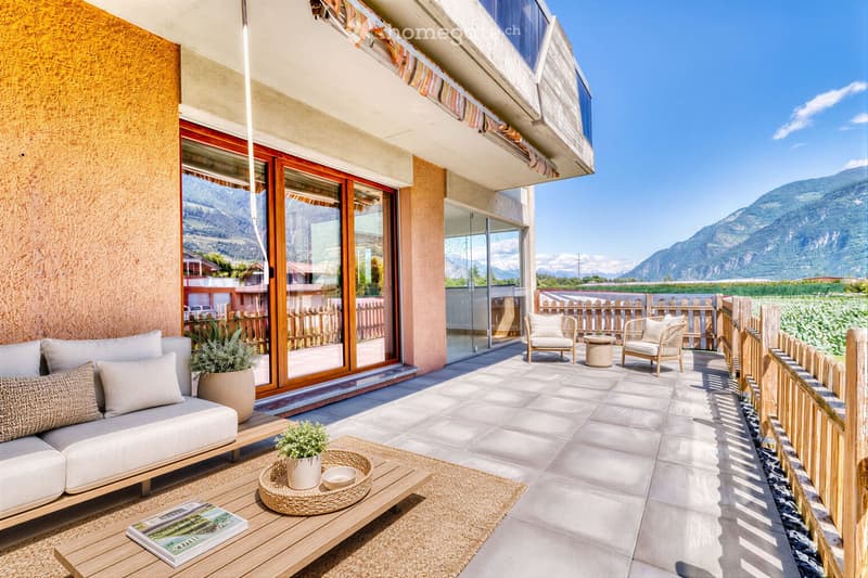 A modern, furnished balcony with a white sofa, wooden coffee table, potted plants, and a scenic mountain view.