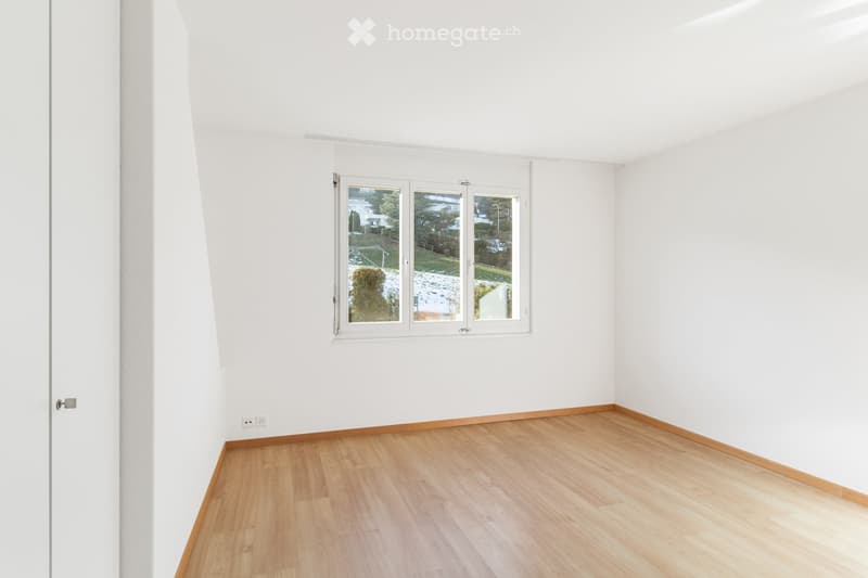 Empty room with white walls, wooden flooring, and a large window offering a view of the outside area.