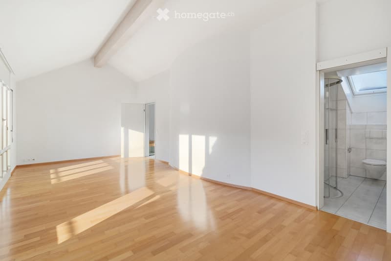 Bright, empty room with wooden flooring and white walls, featuring large windows and a glass-enclosed shower area with a toilet visible in the corner.