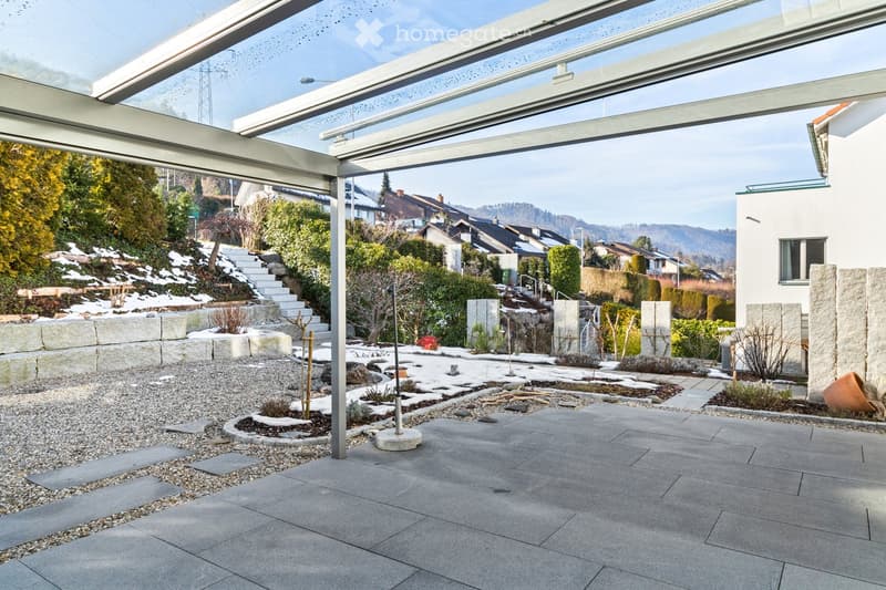 Modern patio with a glass roof structure, stone pathways, gravel areas, small garden beds, and surrounding greenery.