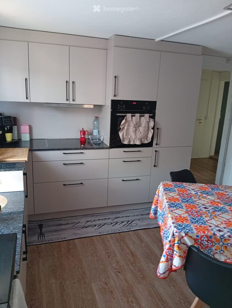 Modern kitchen with white cabinets, stainless steel appliances, and a wooden countertop. Features an oven, coffee maker, and a small dining area with a table and chairs.