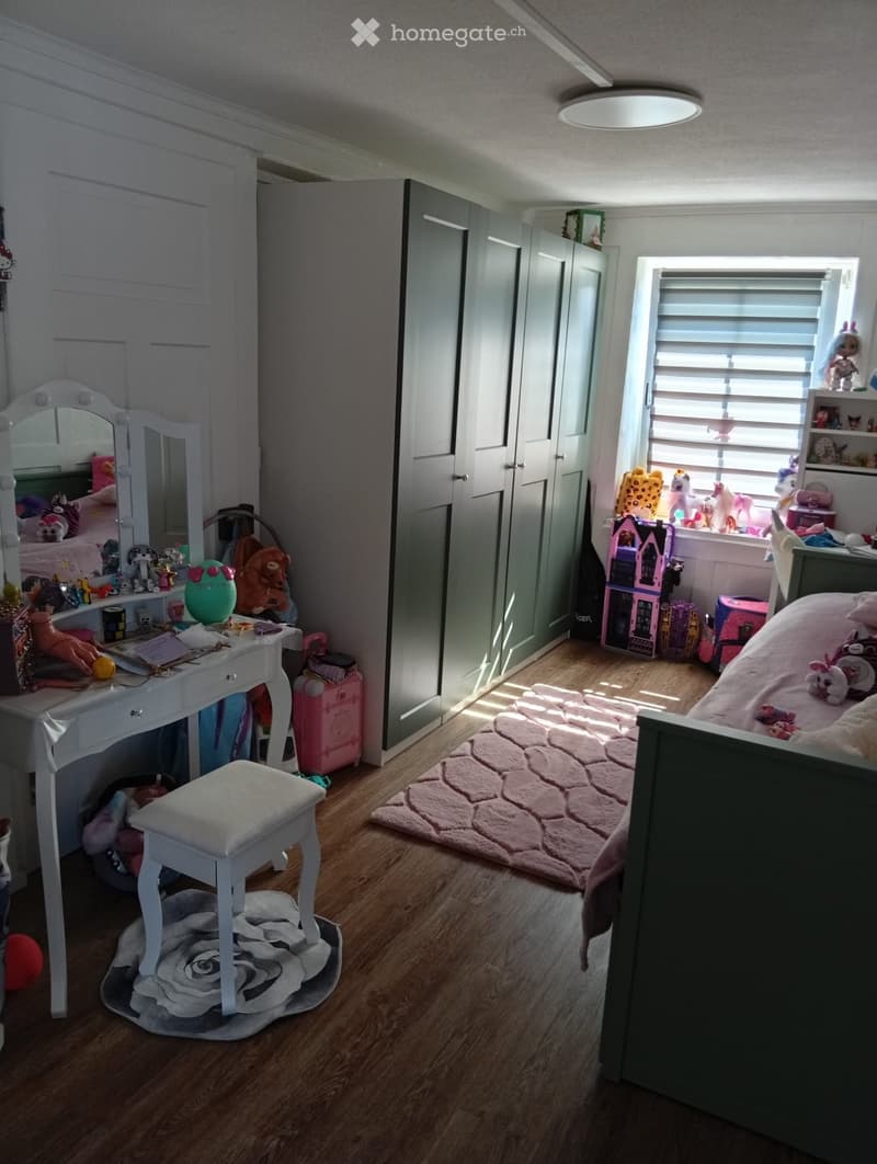 A well-lit bedroom with a bed, a vanity table with a mirror, a stool, and various toys scattered around. The room features wooden flooring and a window with blinds.