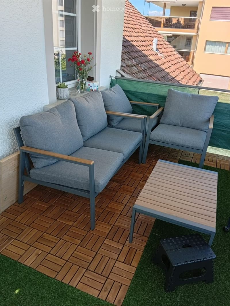 Balcony with wooden decking, grey sofa, armchair, small table, and black stool. Features a small potted plant and vase with red flowers on the windowsill.