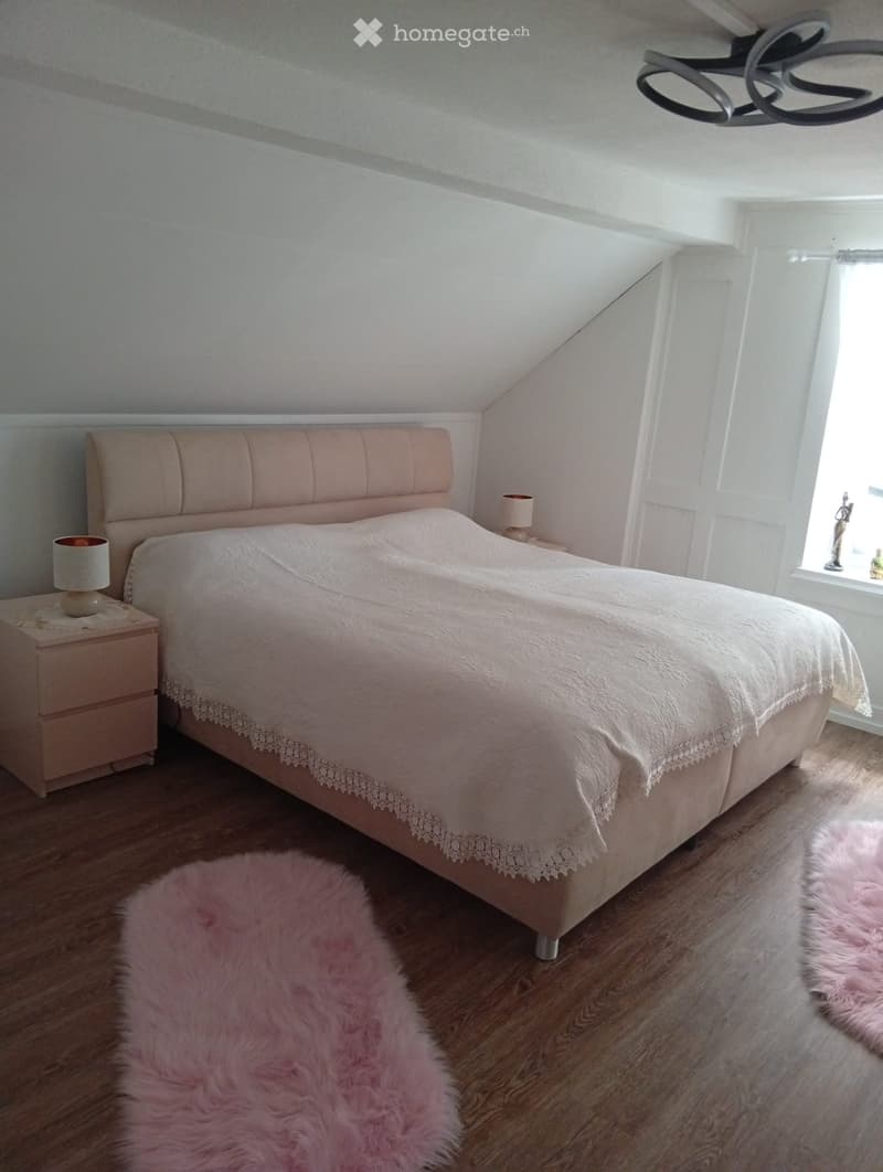 Cozy bedroom with a large bed, white bedding, pink headboard, nightstands with lamps, and pink fur rugs on a wooden floor.