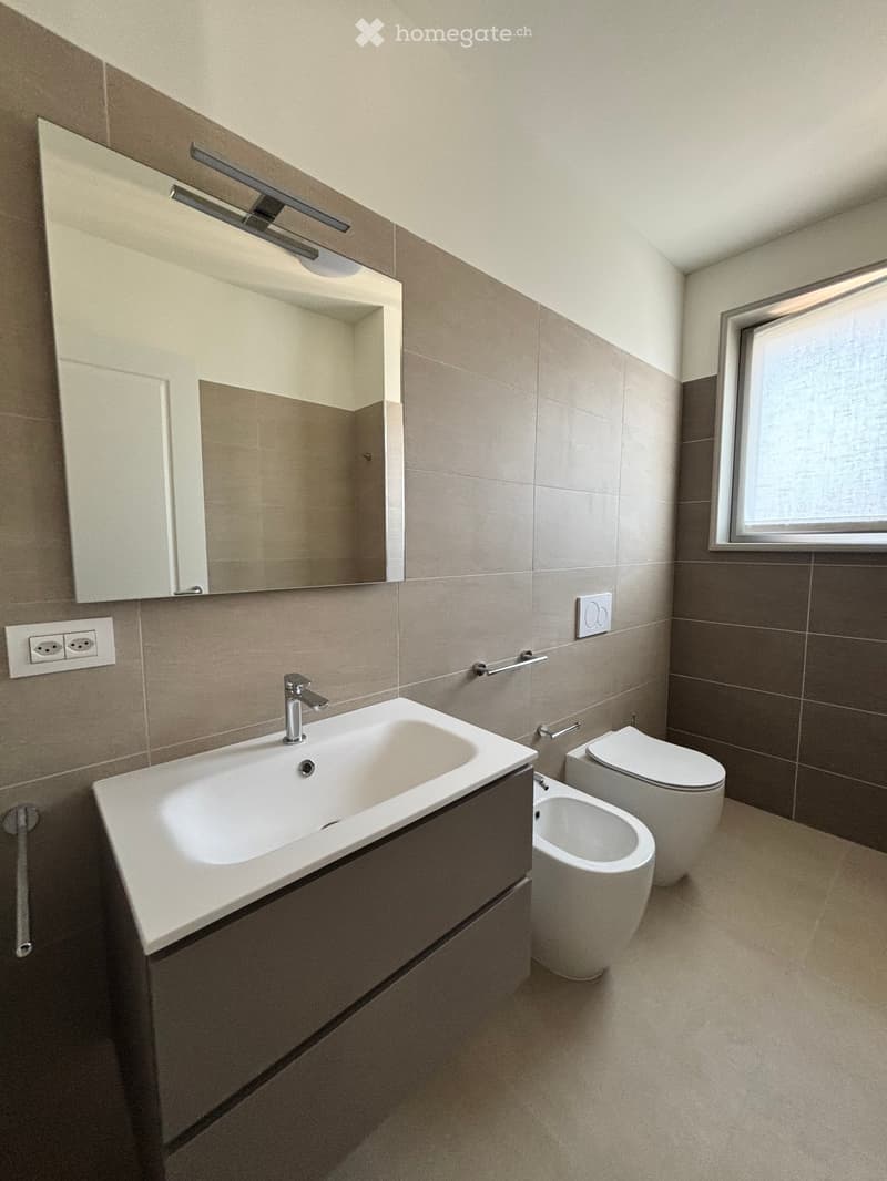 Modern bathroom with beige tiles, a large sink with a chrome faucet, a mirror with built-in lighting, a toilet, and a bidet.