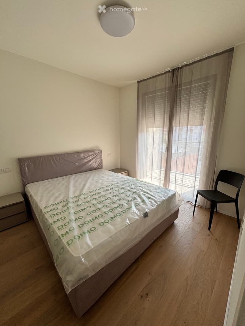Bedroom with a double bed covered in plastic, a nightstand, and a black chair near a large window with sheer curtains.