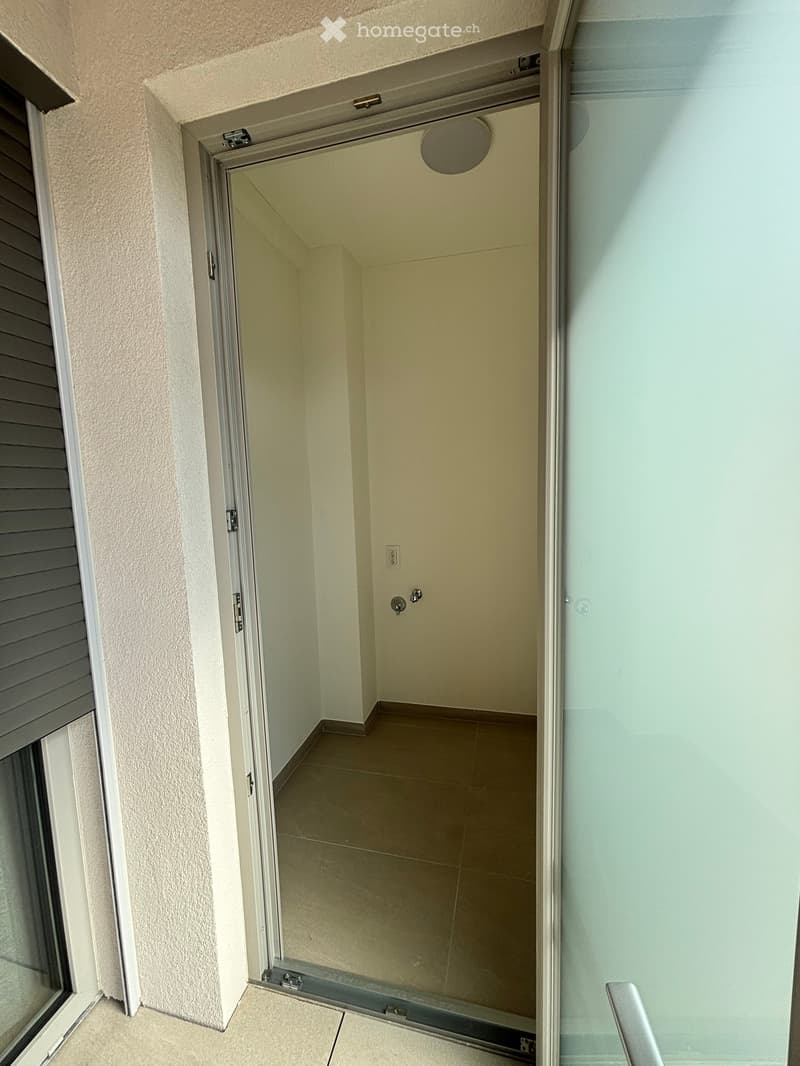 A narrow, empty hallway with beige tiled flooring and light-colored walls, featuring a closed sliding glass door at one end and a partially open door leading to another room.