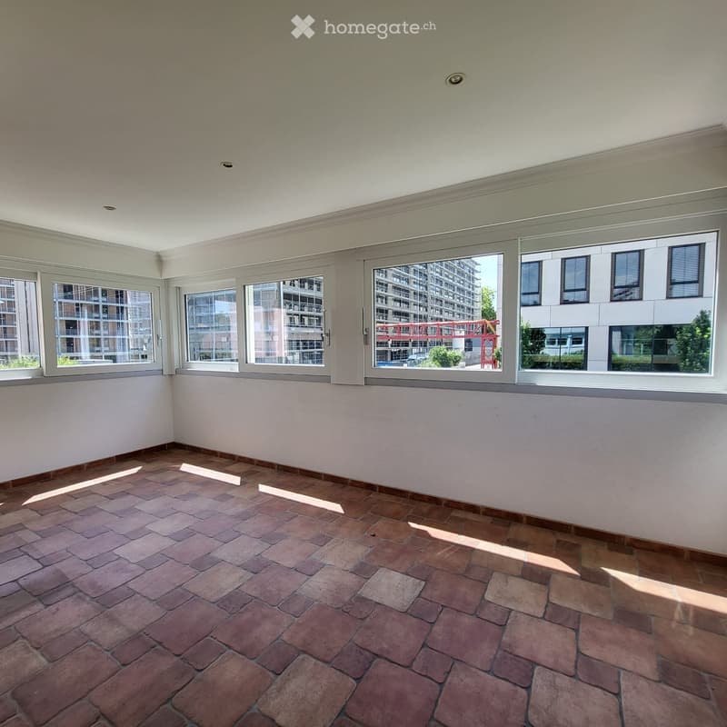 Empty room with brick flooring and large windows offering a view of a modern building and a red structure outside.