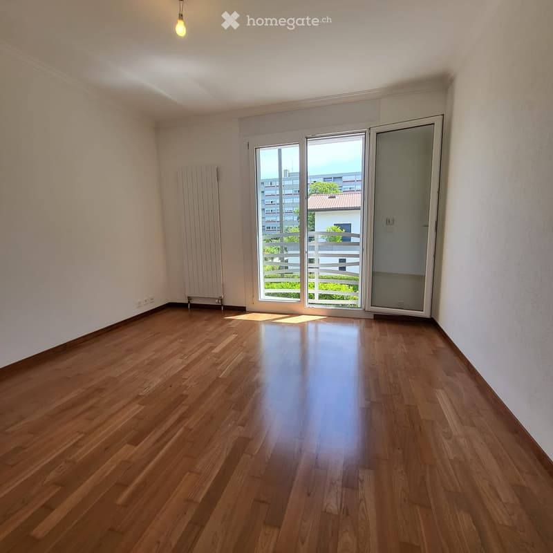 Empty room with wooden flooring, white walls, large window with sliding glass door leading to a balcony, radiator on the left side, and a hanging light fixture.