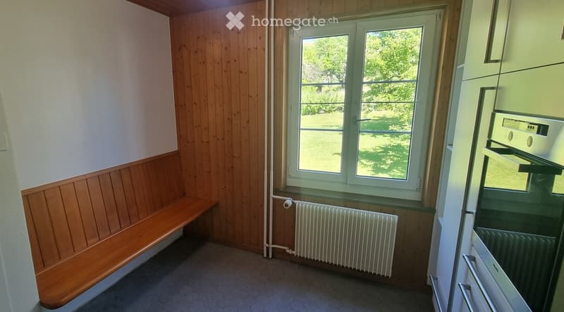 Empty room with wooden paneling, a window with a view of greenery, a radiator, and a built-in bench along one wall.