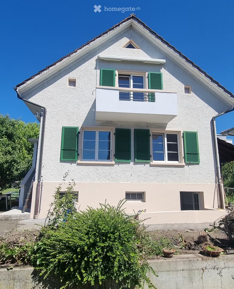 Two-story house with white exterior, green shutters, and a balcony on the upper floor. The house has multiple windows and is surrounded by greenery.