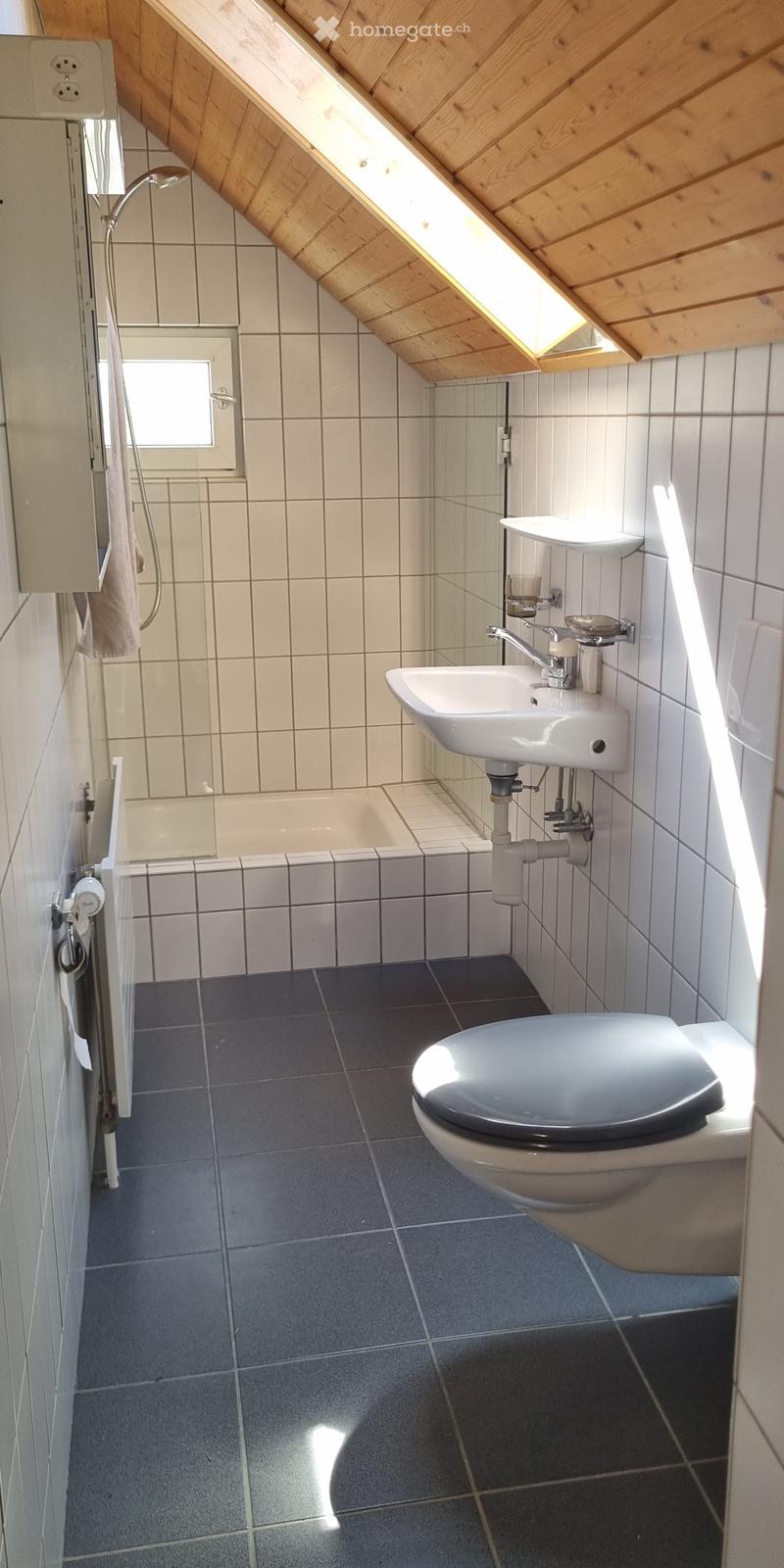 Modern bathroom with a toilet, sink, bathtub, and mirror. The room features grey tiles on the floor and white tiles on the walls, with a wooden slanted ceiling and a small window letting in natural light.
