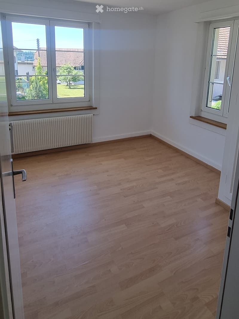 Empty room with light wooden flooring, white walls, and large windows with wooden sills. A radiator is visible under one window.