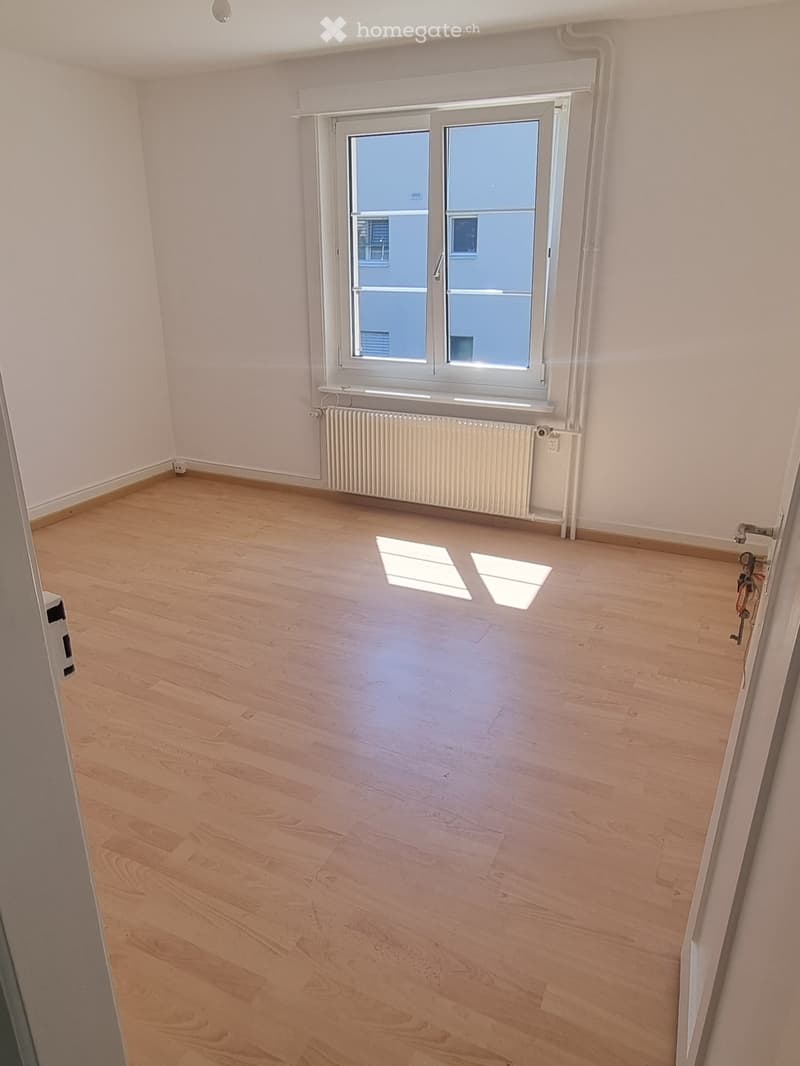 Empty room with light wooden flooring, white walls, and a large window with a radiator below it.