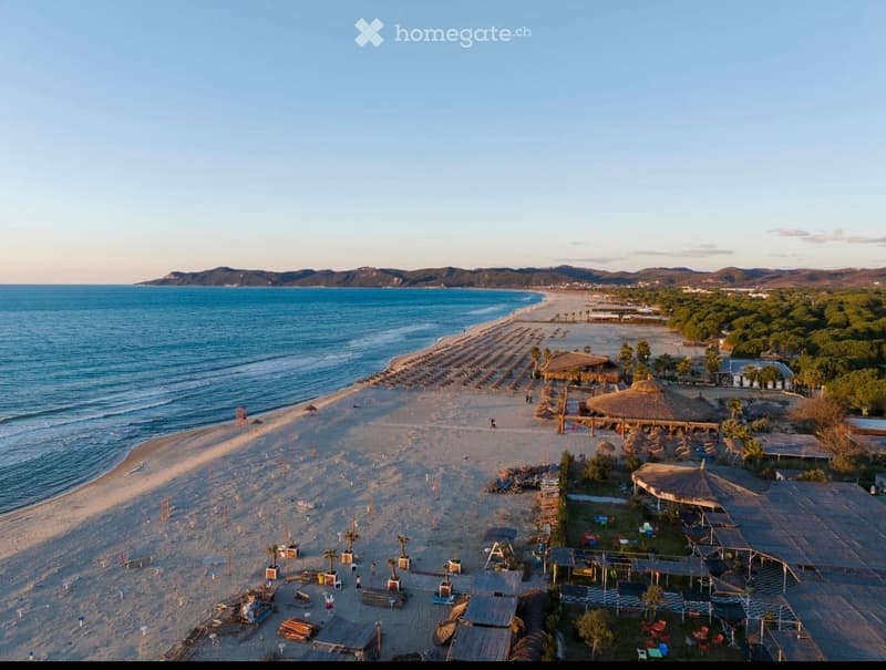 Aerial view of a sandy beach with rows of umbrellas and lounge chairs, surrounded by a wooden deck area with thatched-roof structures and scattered seating, with the sea and distant hills in the background.