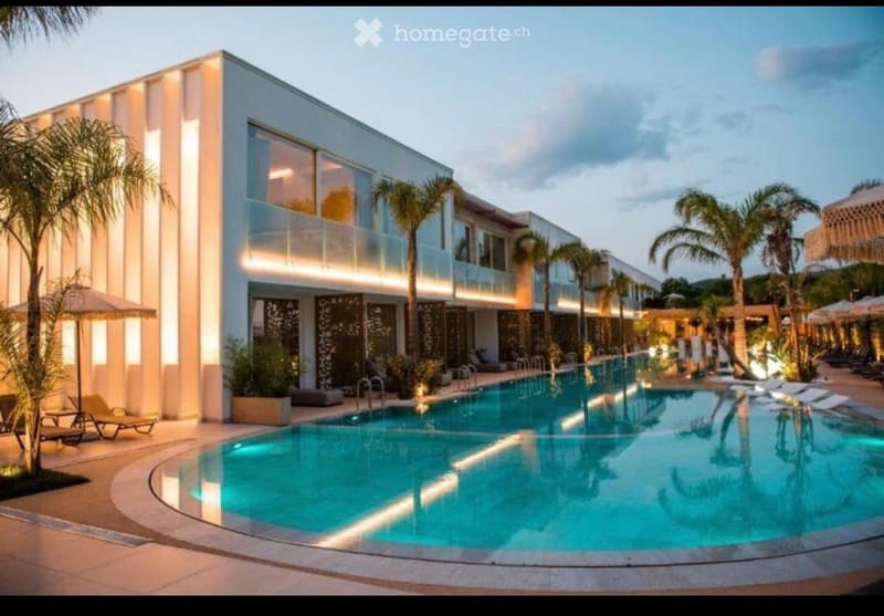 Modern building with a large swimming pool surrounded by lounge chairs, umbrellas, and palm trees.