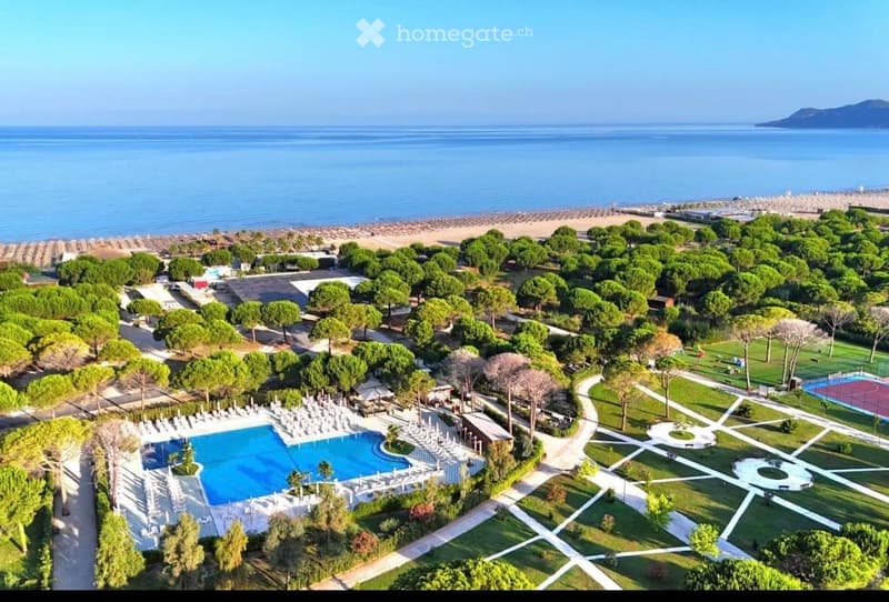Aerial view of a resort with a swimming pool, lounge chairs, trees, pathways, and a view of the sea and beach.