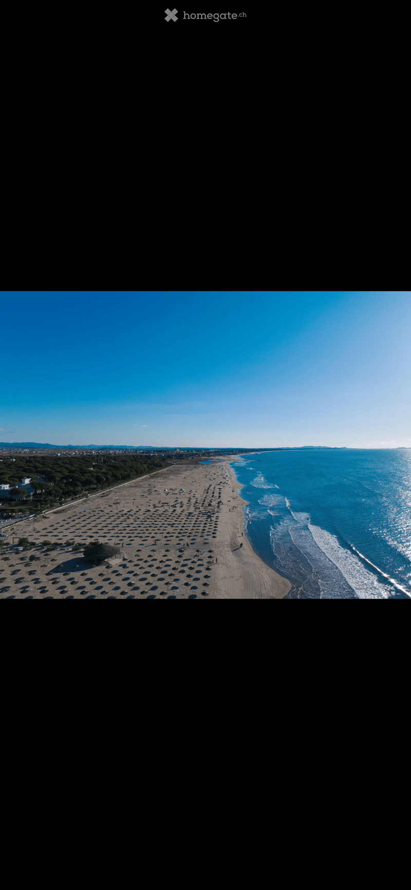 Aerial view of a sandy beach lined with numerous umbrellas and lounge chairs, with the sea visible on the right side and distant land on the left.