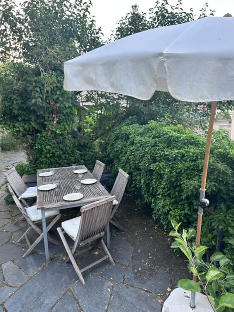 Outdoor patio area with a wooden dining table, four chairs, and a large umbrella providing shade. The setting includes greenery and a stone-paved floor.
