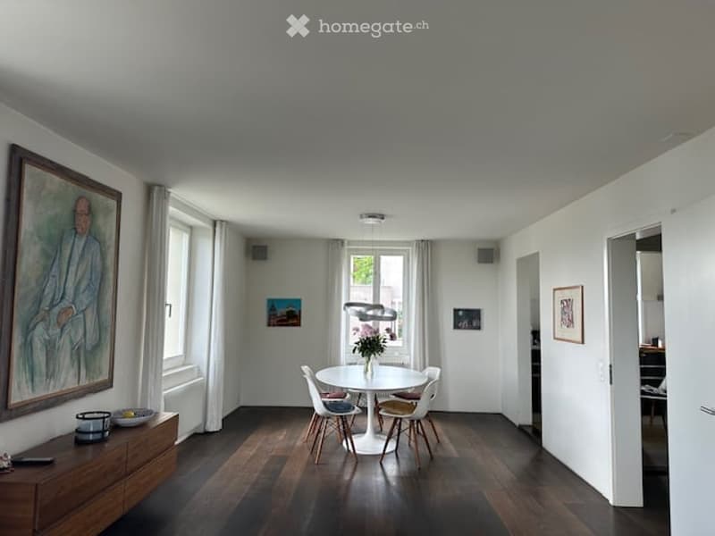 Dining room with a white round table, white chairs, wooden floor, white walls, a painting on the wall, a window with white curtains, a radiator, and a wooden cabinet with a bowl and a candle on top.