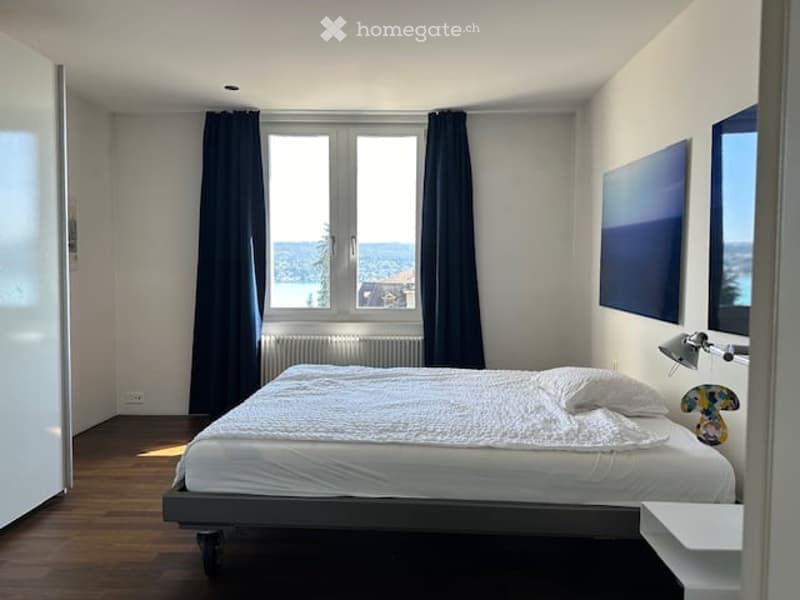 white walls, wooden floor, large window with view, bed with white sheets, white pillows, radiator, lamp, artwork