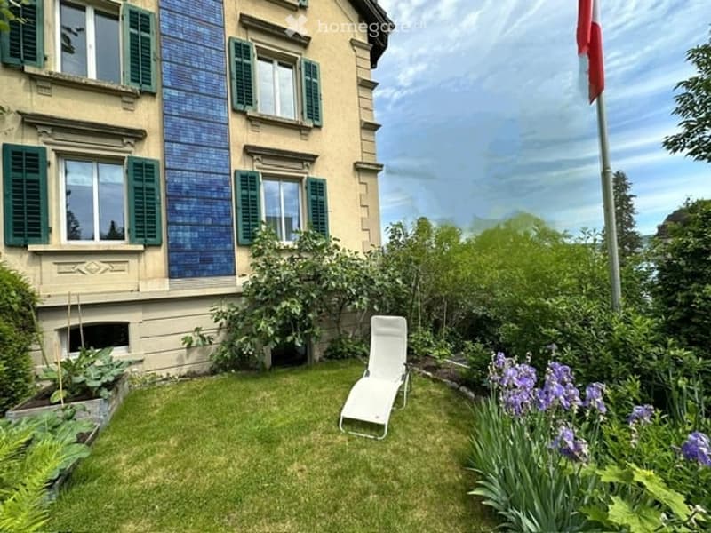 A garden with a white lounge chair, solar panels on the wall, and a flagpole with a red flag.