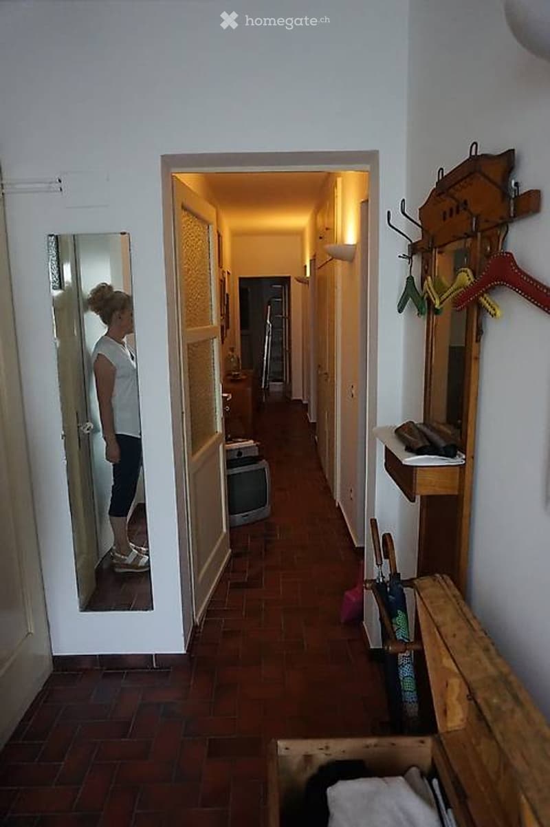 A narrow hallway with wooden flooring and white walls, featuring a coat rack with several hangers, a small shelf with items, and a partially open door leading to another room.