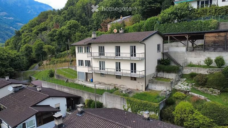 A two-story residential building with a beige facade, multiple windows, and balconies, surrounded by greenery and situated in a hilly area with visible mountains in the background.