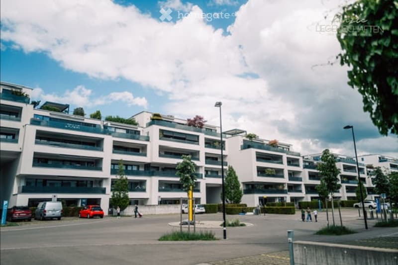 Modern multi-story residential building with balconies, surrounded by a paved area with parked cars and a few pedestrians.