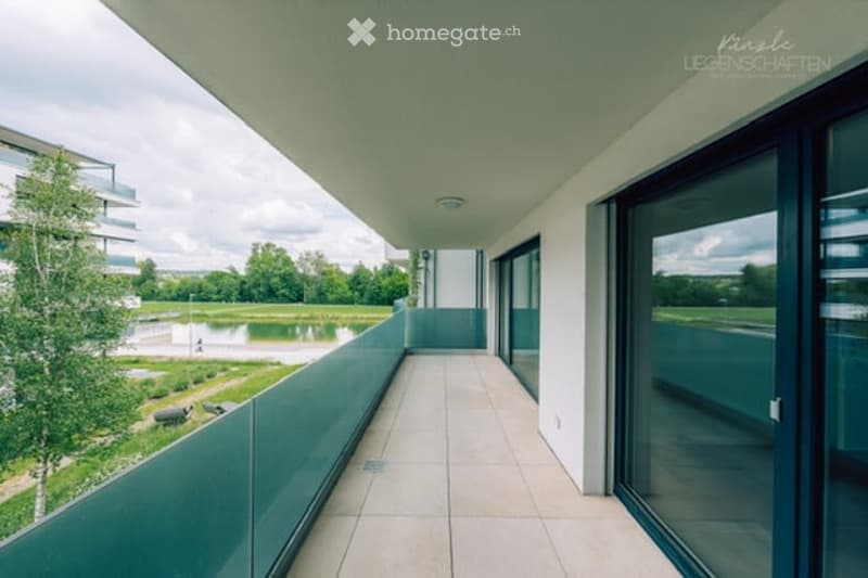 Modern balcony with large glass sliding doors, tiled floor, and a view of a landscaped area with water and greenery.