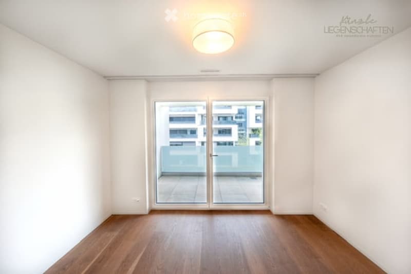 Empty room with wooden flooring, white walls, large sliding glass door leading to a balcony, and a ceiling light fixture.