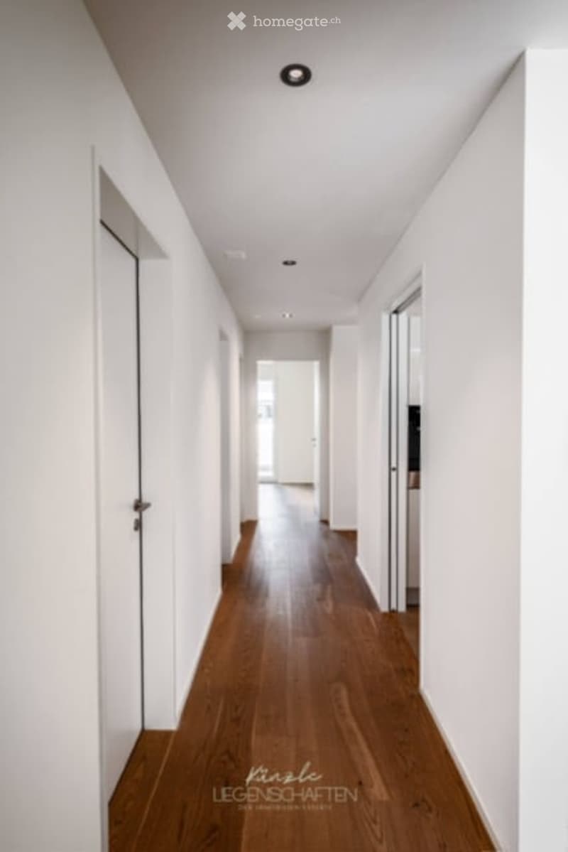 Clean, modern hallway with light wooden flooring, white walls, and recessed ceiling lighting. Doors on both sides lead to other rooms.