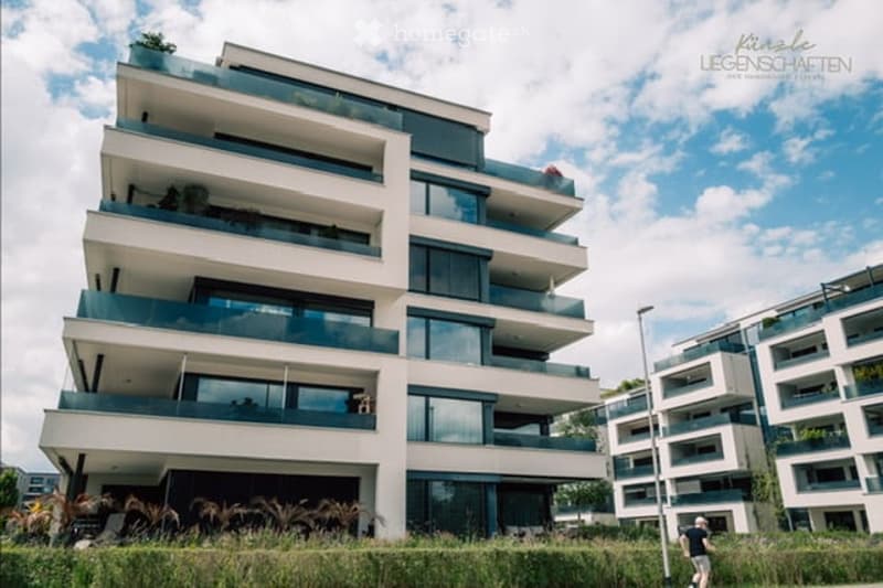 Modern multi-story residential building with balconies and large windows, surrounded by greenery.