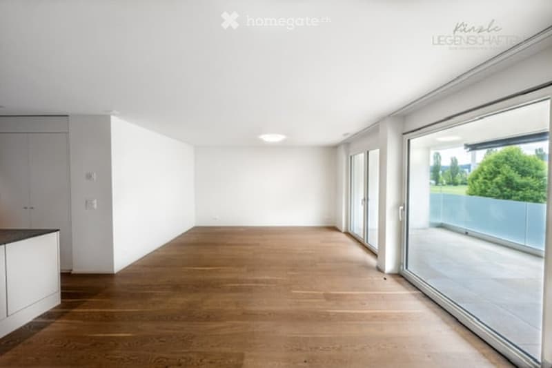 Bright, empty room with wooden flooring, white walls, large windows, and a view of greenery outside.