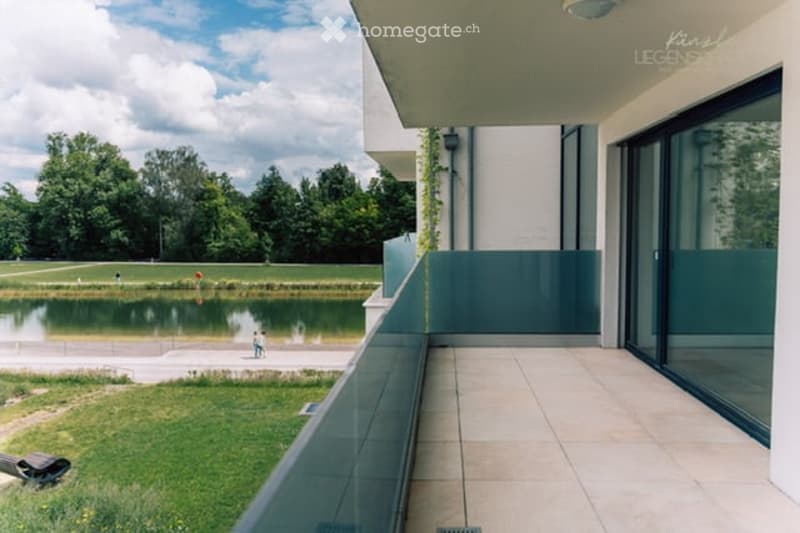 Modern balcony with large glass railings, tiled floor, overlooking a serene lake and green park area.