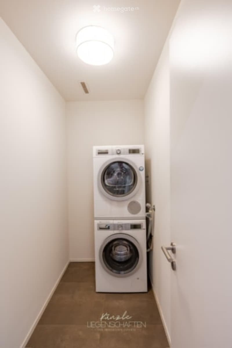 Small, clean laundry area with a stacked washing machine and dryer unit, white walls, and a ceiling light.