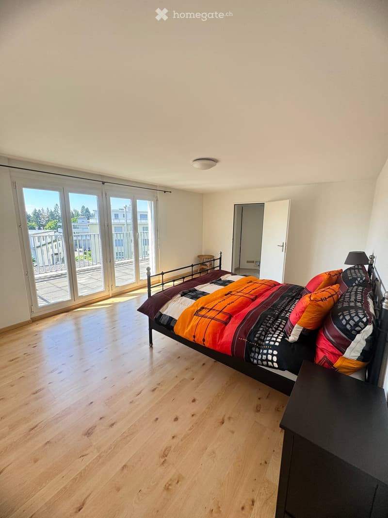 Bright bedroom with wooden flooring, large sliding glass doors leading to a balcony, a bed with colorful bedding, a nightstand, and a small stool near the bed.
