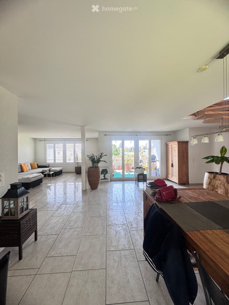 Bright living room with tiled floor, large windows, sliding glass doors leading to a patio with a swimming pool, sofa, coffee table, dining table, chairs, plants, and a cabinet.