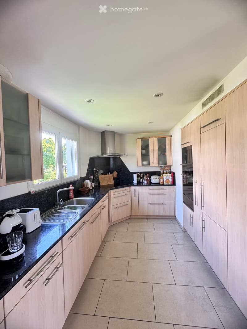Modern kitchen with light wood cabinets, black countertops, stainless steel sink, and various appliances including a toaster and oven. The space is well-lit with large windows and has a clean, organized layout.