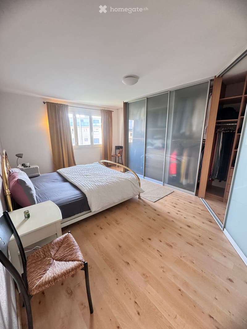 Modern bedroom with wooden flooring, large window with curtains, sliding glass door leading to balcony, bed with decorative pillows, nightstand, and a woven chair.