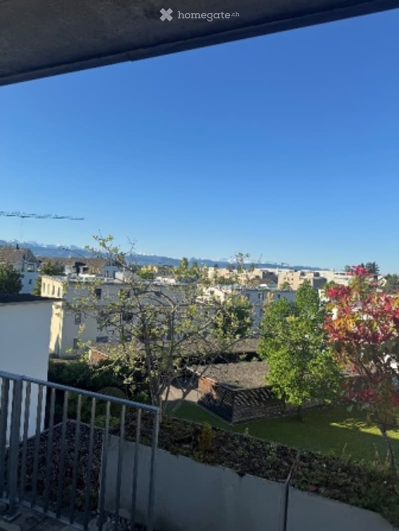 Balcony with metal railing, overlooking a cityscape with buildings, trees, and distant mountains under a clear blue sky.