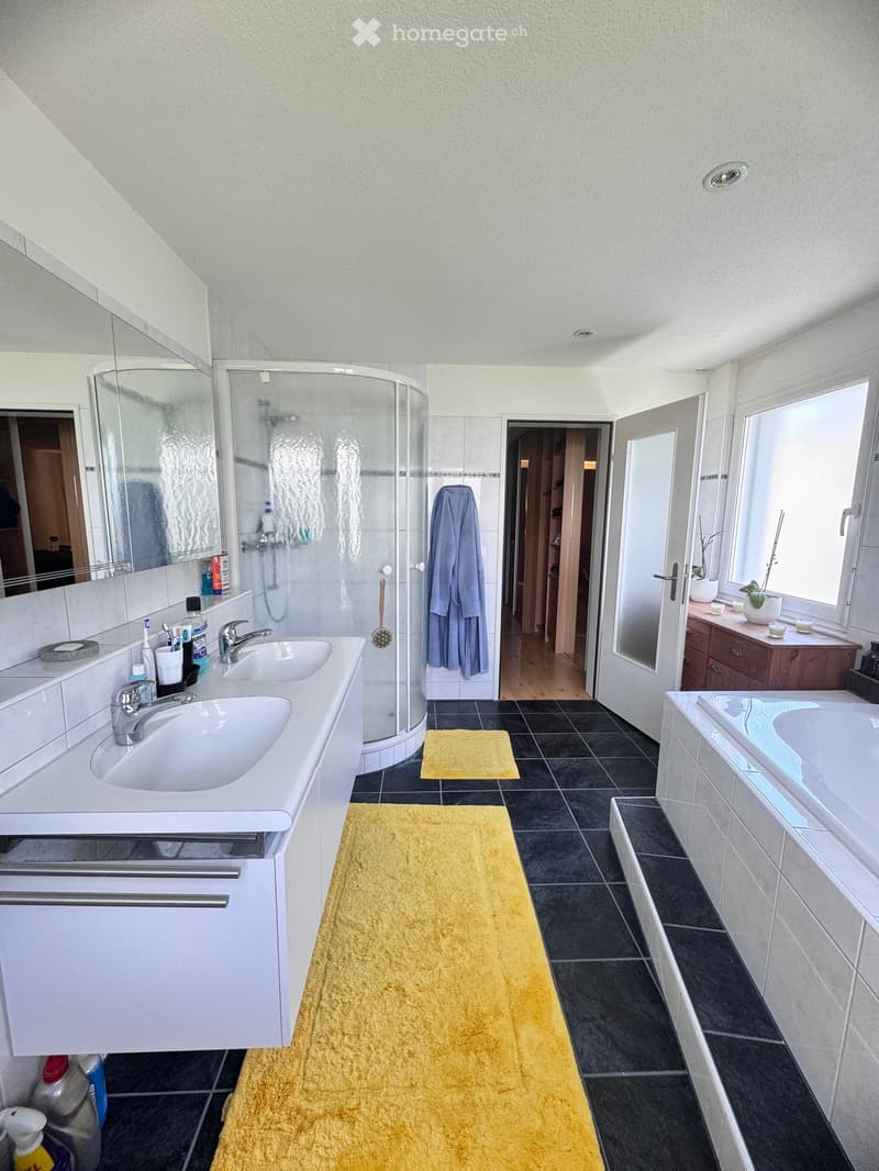 Modern bathroom with double sink, bathtub, shower, and large mirror. Features black tiled floor, white walls, and yellow rugs.