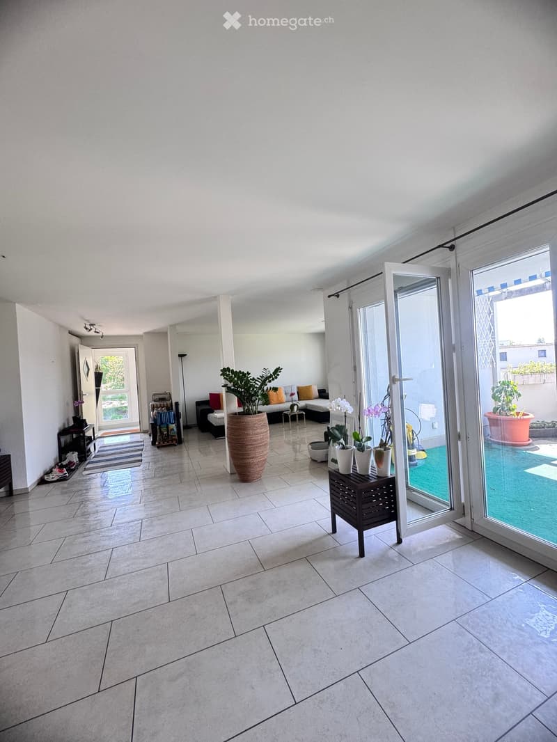 Bright, open living space with tiled flooring, large sliding glass doors leading to a patio, and several pieces of furniture including a sofa, side tables, and potted plants.
